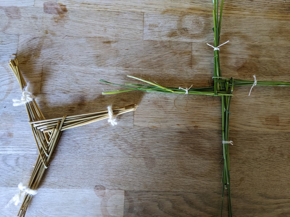 A triskele Brigid's cross, dried out and light brown, woven in 2025 with Irish river rushes and tied with white yarn. Next to it is a freshly woven four-legged Brigid's cross, bright green, woven with grasses picked from the unkempt patch of earth on my street corner.