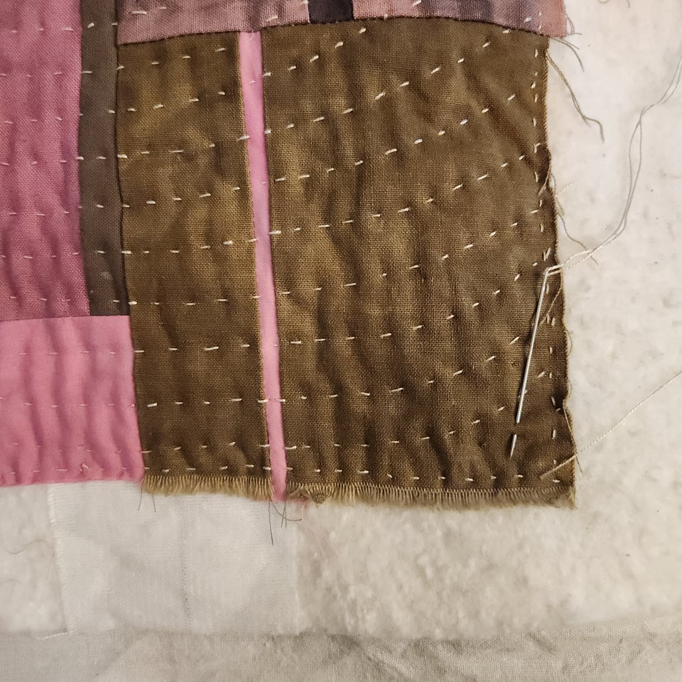 Close up photo of the corner of a patchwork quilt held together with white stitches. The needle used to stitch the quilt together is in the corner of the fabric. Quilt batting is visible around the patchwork. The fabric colors are olive green and several shades of pink.