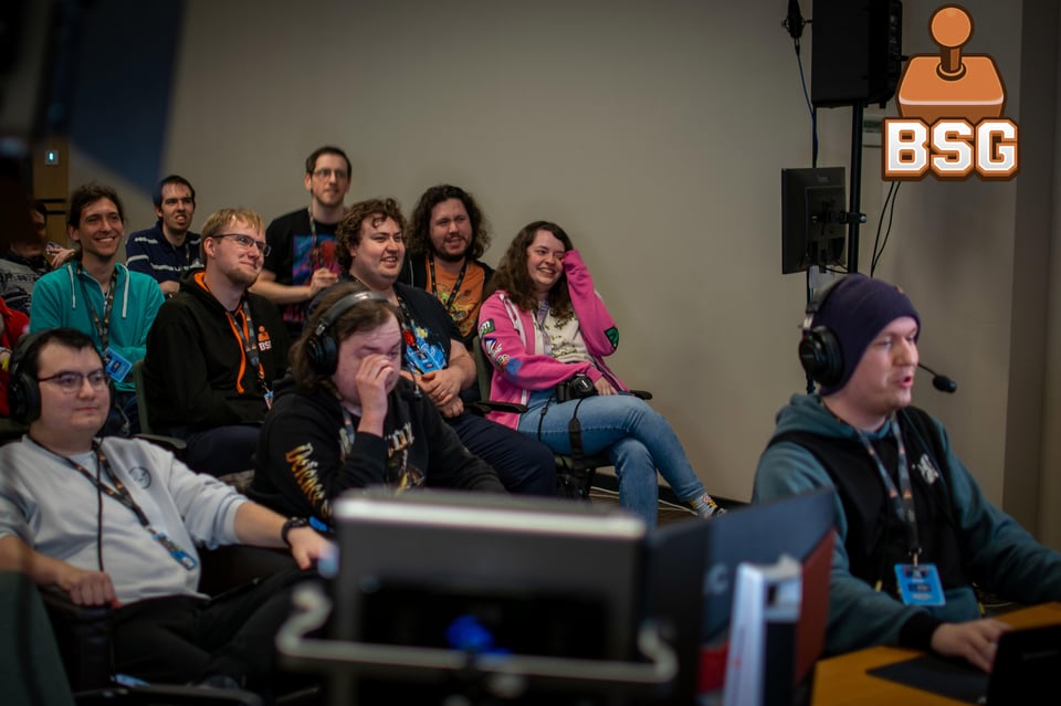 A group of people, smiling, laughing, watch a speedrunner in headphones at the head of a small room play through a game.