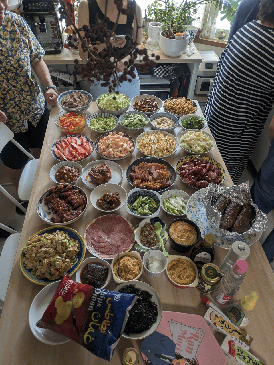 A table full of plates and bowls of ingredients, including hummus, powdered peanut, sliced meat, green onions, cilantro, shrimp, eggs, and more.