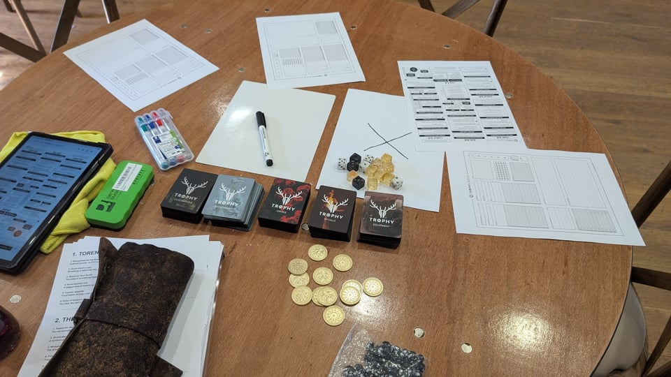 a tabletop roleplaying game, Trophy Gold, laid out on a table in a castle; gold hunt tokens, character creation cards, light, dark and gold dice and character sheets, pens, etc...