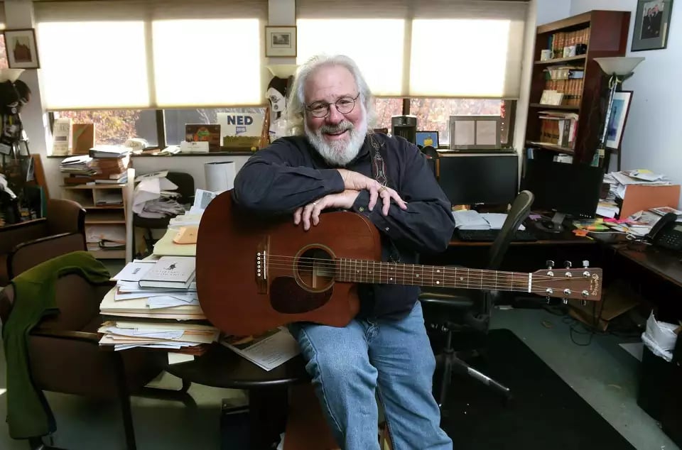 A man with a graying beard and glasses sits on teh corner of a desk piled with papers, his right hand resting on his left hand and holding a Martin acoustic guitar.