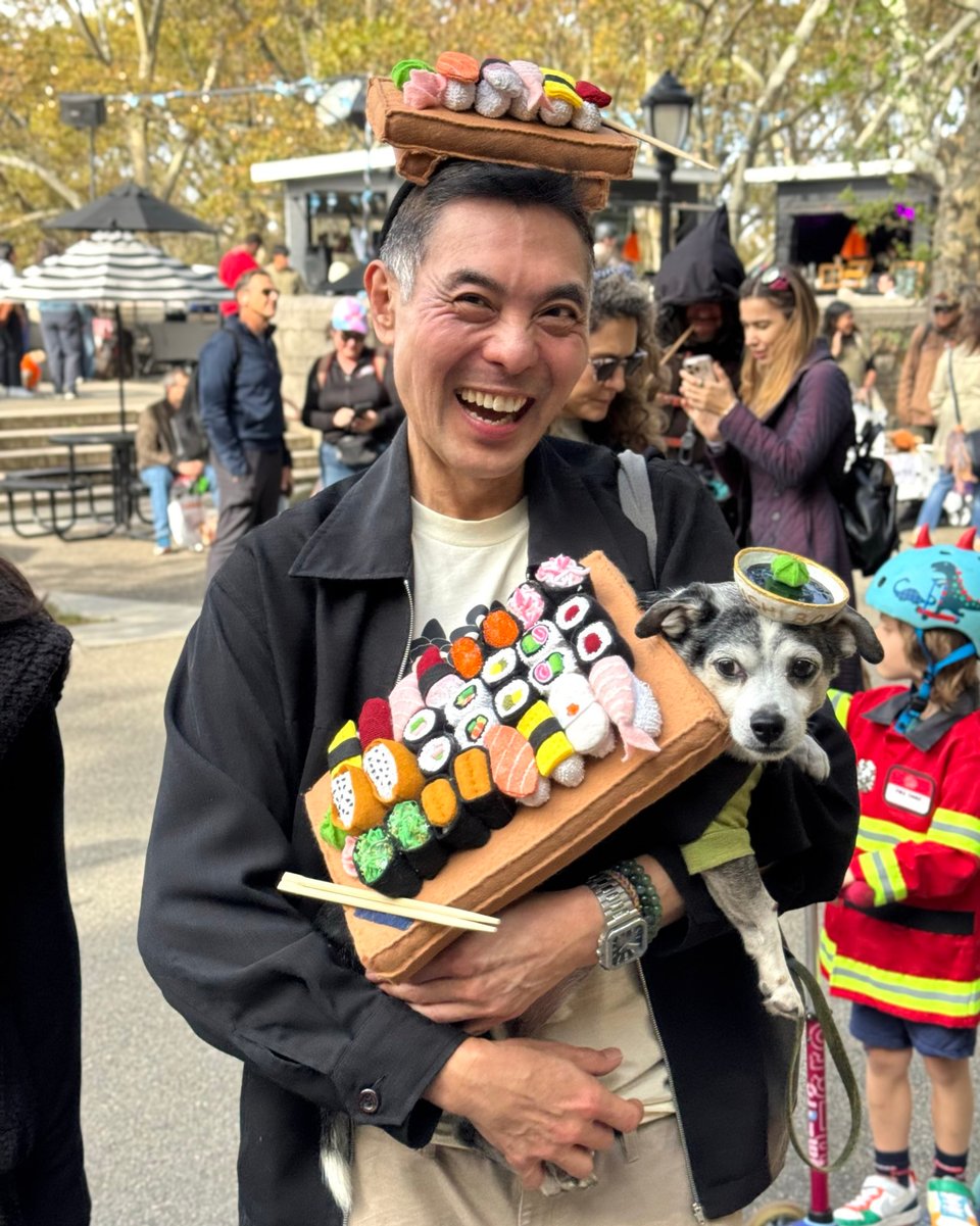 A smiling man holds a dog wearing an elaborate sushi costume. The man wears a matching hat