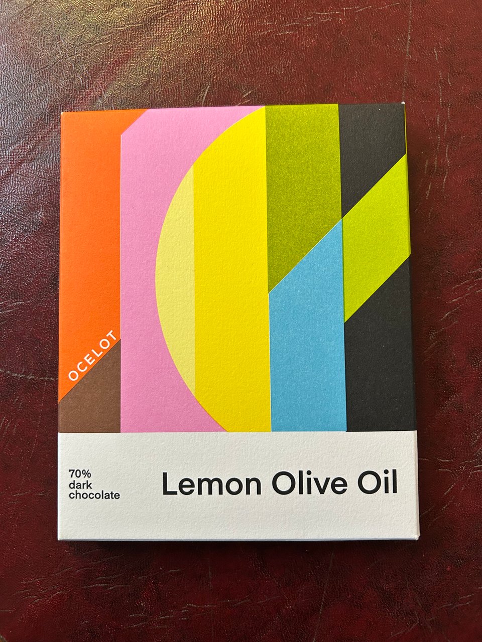 A bar of Ocelot lemon olive oil chocolate with its colourful packaging is sitting on a dark red leather topped table.