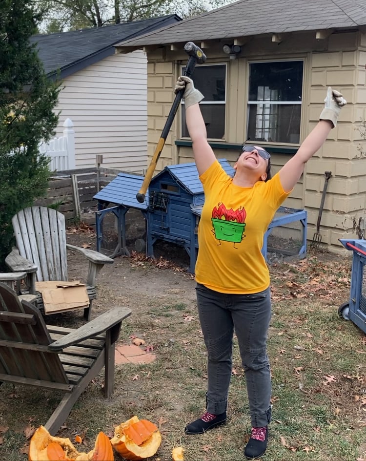 Woman wearing dumpster fire t-shirt with arms in the air after destroying a pumpking in her backyard.