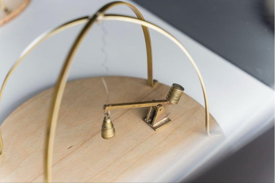 a close up of a brass mechanism with a weight pulling a wiggly wire, on a wooden base