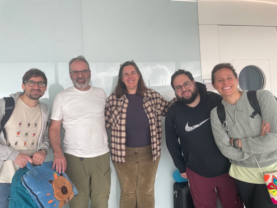 Five people standing in a line against a white wall all smiling at the camera.