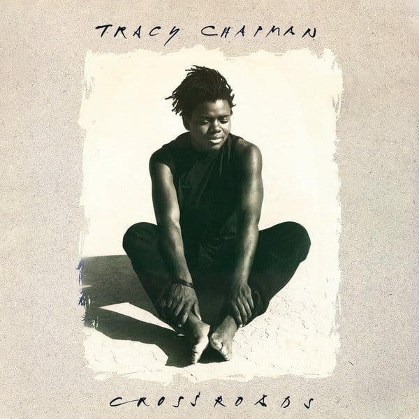 A slightly sepia-toned photo of Tracy Chapman, a young Black woman with short dreadlocks, sittng on sand. She holds her ankles. She wears a sleeveless black shirt, black pants, and is barefoot. Her eyes are closed, as if thinking hard or maybe just really really experiencing the sun reflecting on her.