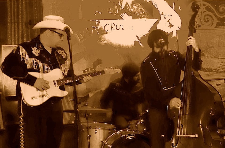 A guitarist wearing a white cowboy hat and fringe snapfront shirt plays a white Fender Telecaster to the right of man seated and playing drums. To the drummer's left, a man in a black cowboy shirt plays an upright bass.