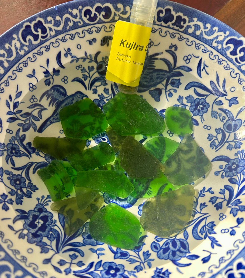 A blue and white china bowl with pieces of green sea glass immersed in water. There's also a small vial of perfume with a yellow label on. Image by Rowan Ambrose.