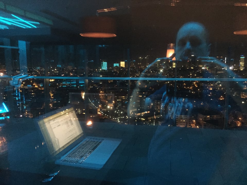 A picture taken from a tall building, at night, with a huge sprawling city beyond. In the reflection of the glass, a laptop can be seen on the table, and the ghostly reflection of a man taking a picture with a phone.