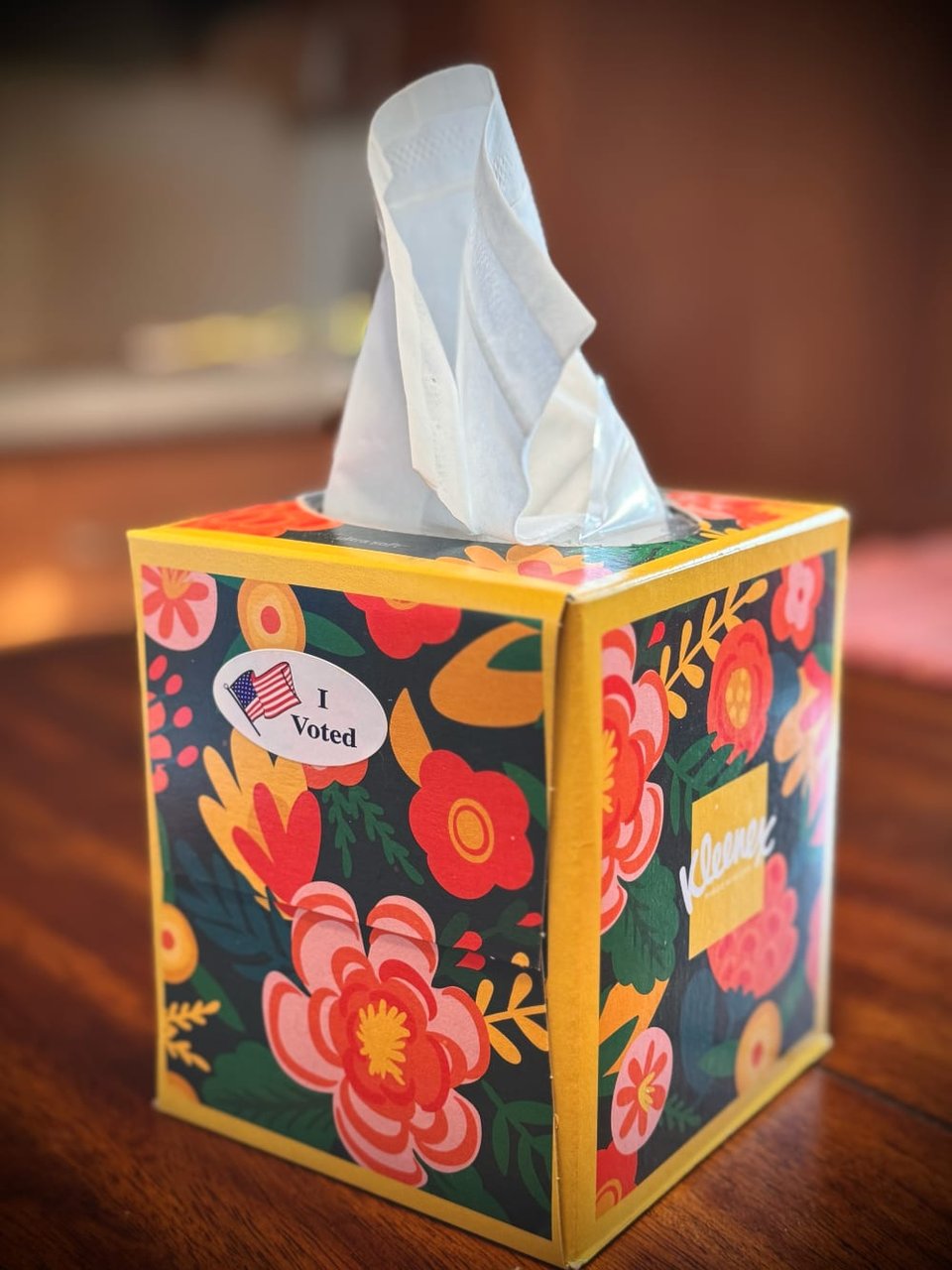 A colorful floral design decorates a tissue box with a tissue popping out of the top of the box. One one side facing you is an "I Voted" sticker with an American flag. On another side of the box the Kleenex logo is visible.