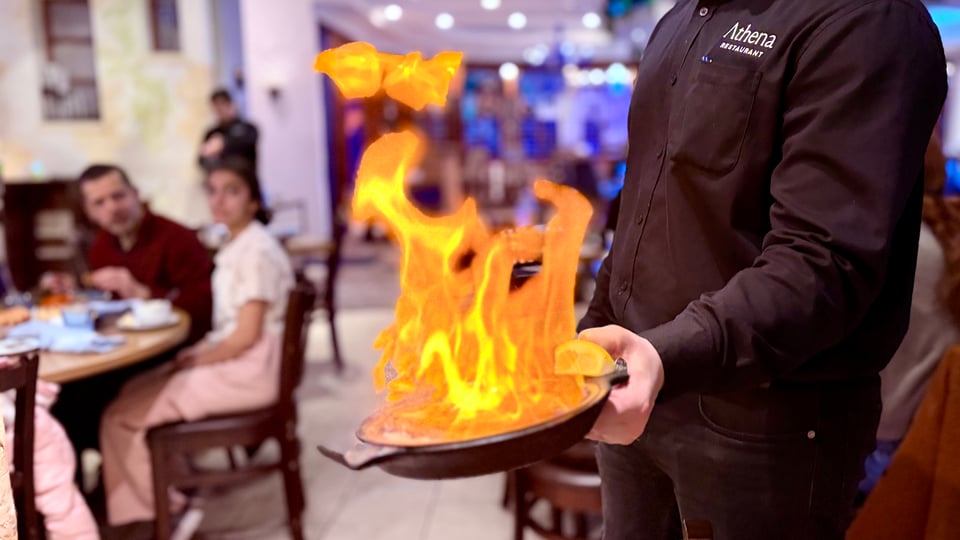 Greek restaurant: in the center, a waiter (no head in the frame) is holding a skillet with a flaming saganaki. On the left, blurred in the background, two customers sitting at a different table observe the scene