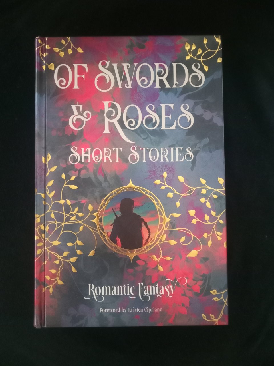 A copy of Of Swords and Roses, an anthology of romantasy short stories from Flame Tree Press. The cover has vines in gold leaf and the image of a warrior with a long braid in a medallion at the center. The edges, which you can’t see here, are sprayed with a pink floral design.