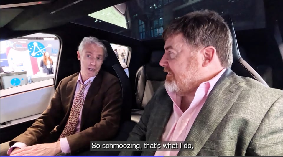 Screencap of two white men inside a parked Cybertruck, doing an interview. The man farther from the camera is subtitled saying, “So schmoozing, that’s what I do.”