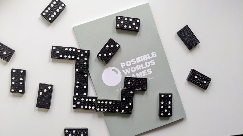 On a white surface lies a zine, a chain of dominos, and scattered dominos. The zine is backside up: there is a logo there of an orb and the words 'Possible Worlds Games'.