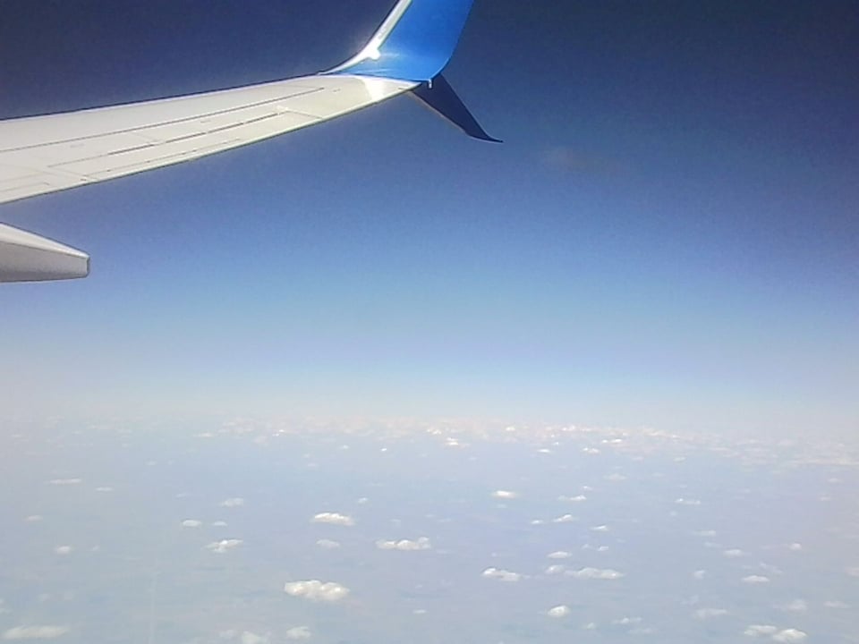 low-res image of the view from a plane window, the edge of the plane wing over a deep blue sky fading out into distant landscapes