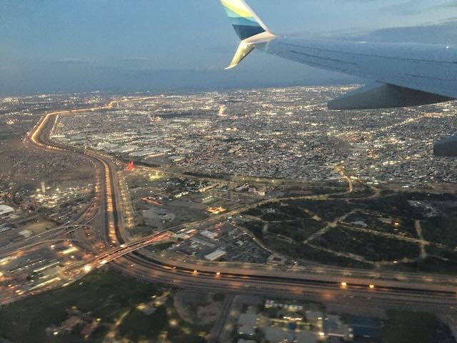 A plane wing flying over a densely built landscape at dusk, with a brightly lit stripe of road and other barriers cutting from one side of the photo to the other.