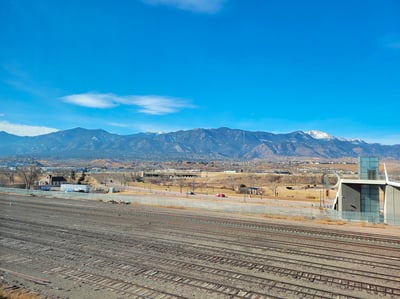 Mountains rise up in the distance under a clear blue sky, with one snow-covered mountain on the right. In the foreground are freight train tracks.