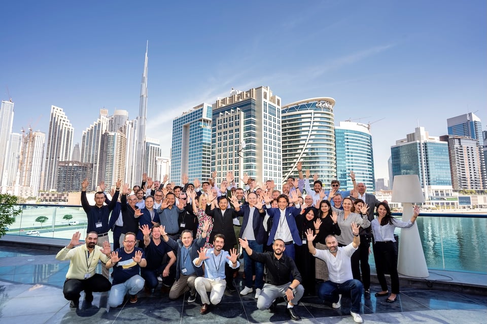 A large group of people in business clothes wave together to the camera, beneath the Dubai skyline.