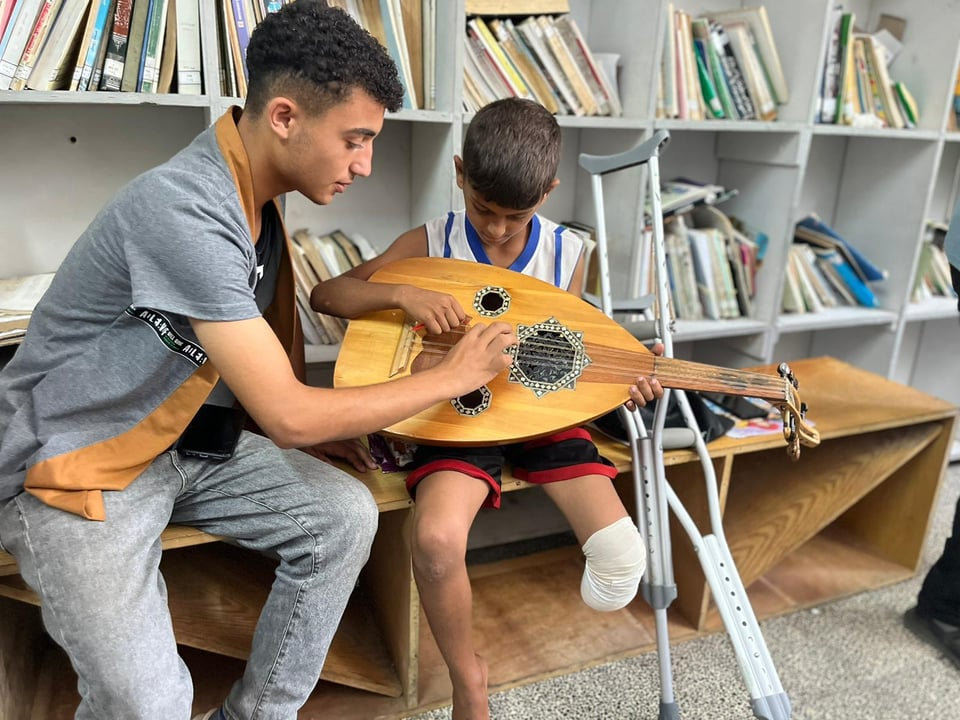A teenage boy with a young boy who has a bandage on his amputated leg, playing an oud. A pair of crutches lies nearby