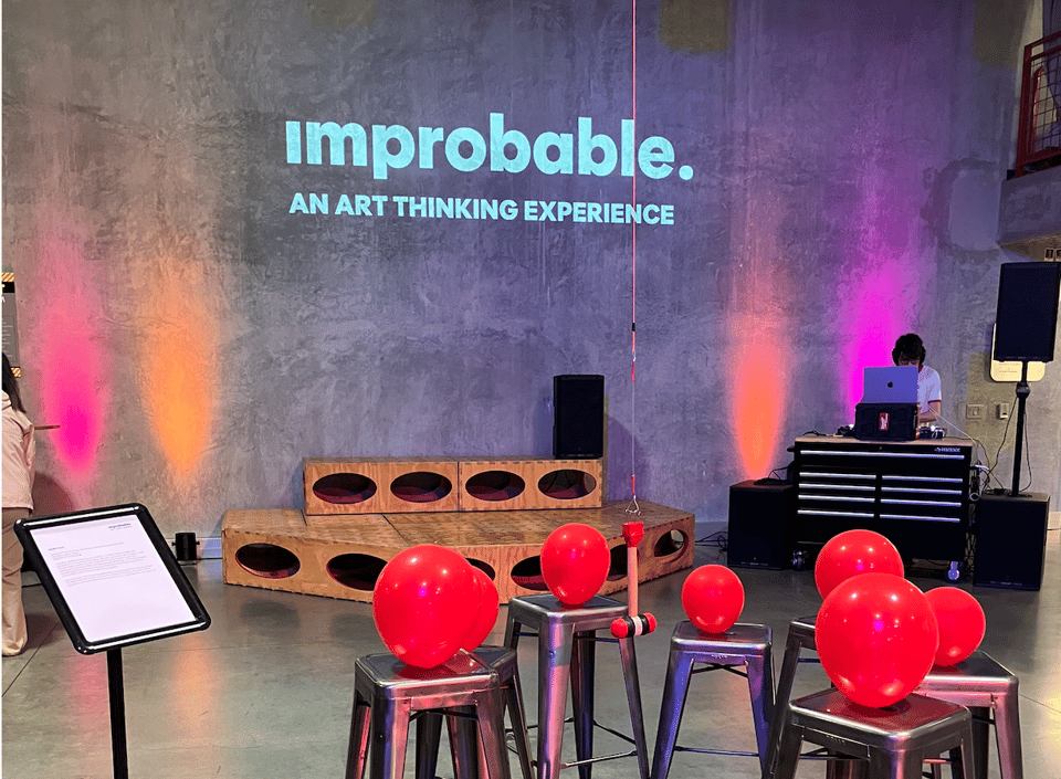 balloons tied to stools sit in front of a colorful wall that reads "improbable. an art thinking experience"