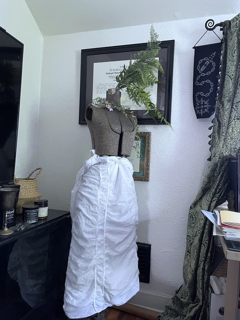 A grey dressmaker's dummy with white, semi draped and gathered cloth wrapped around the lower half. There are plastic ferns sticking out of the neck.