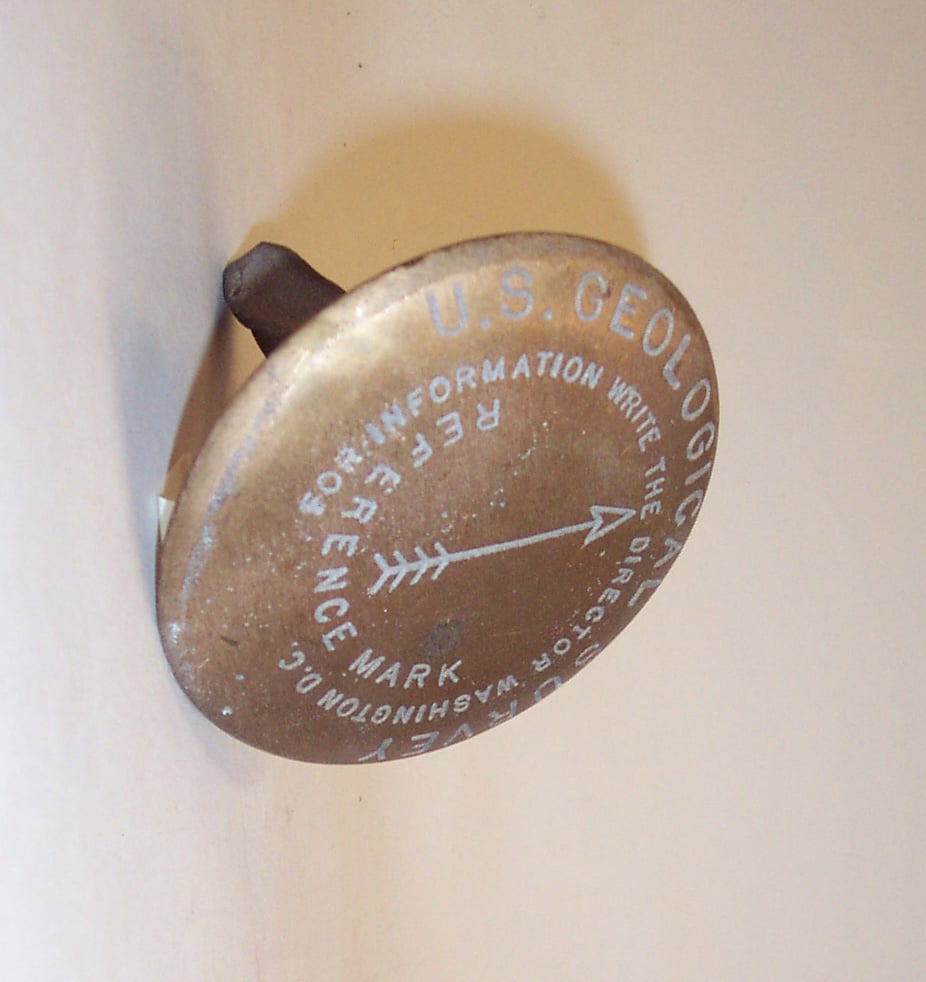 Photograph of a vintage brass U.S. Geological Survey marker, shown at an angle against a light background. Text reading 'U.S. GEOLOGICAL SURVEY' and 'REFERENCE MARK' are visible on its circular face, with a needle or pointer indicator.