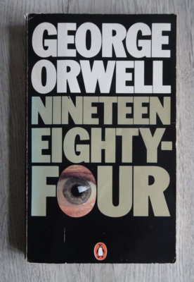 a book, the cover is black, the top third of the book has bold white writing George Orwell, the bottom two thirds has grey bold writing with the O of the four showing a large eye