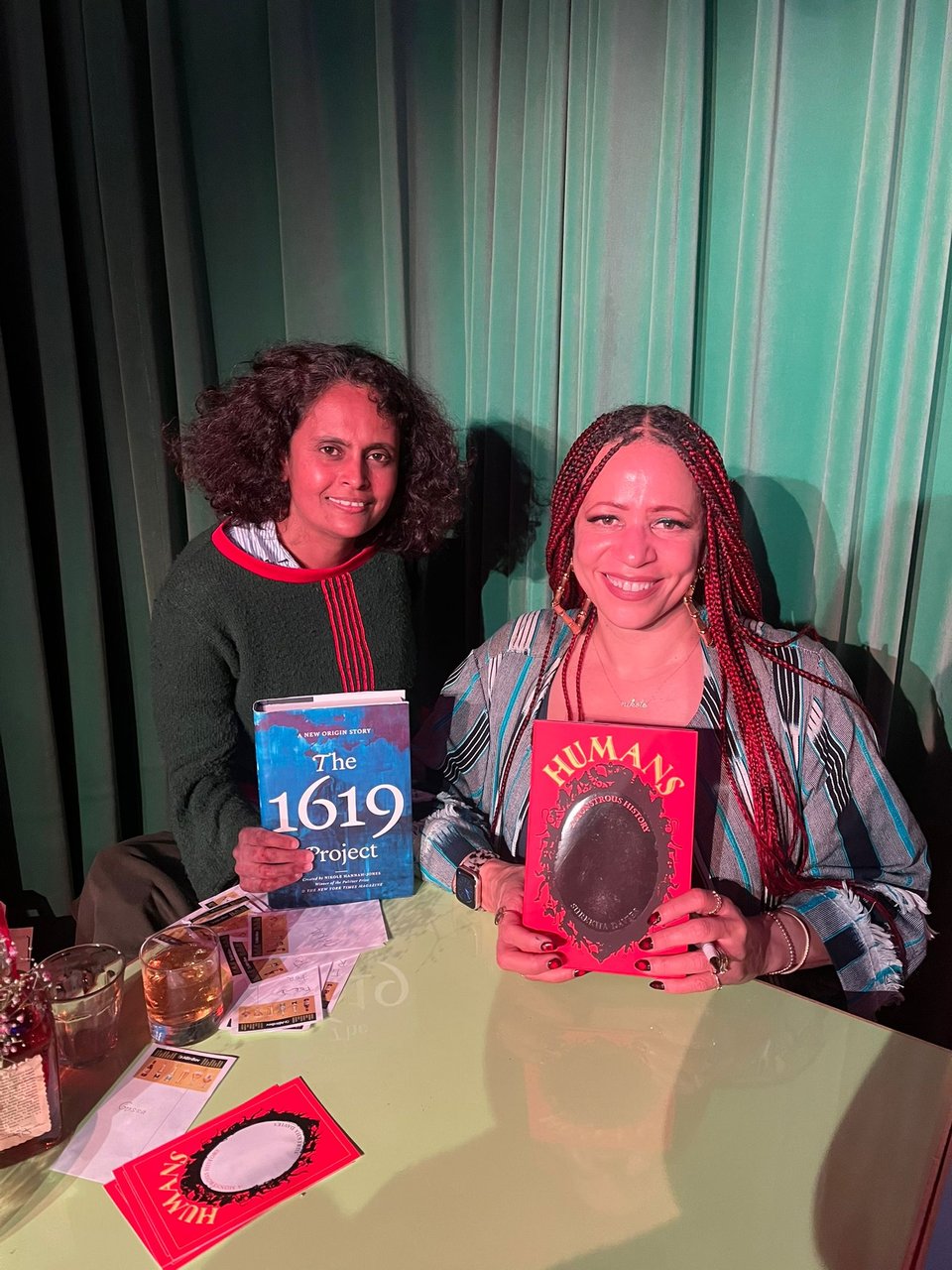 Me and the amazing Nikole Hannah-Jones behind a desk, with a green curtain behind us, in pinkish light, smiling. Nikole is holding up the bright red and silver HUMANS, and I'm holding up Nikole's blue and white THE 1619 PROJECT. Nikole is seated; I'm standing but crouching down the left.