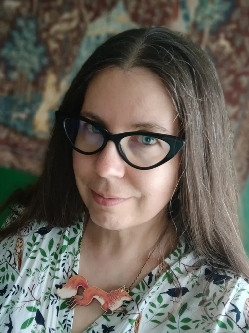 headshot of Deva Fagan, a woman with pale skin, blue eyes, long brown hair, and black cat-eye glasses. she's wearing a blouse printed with birds and leaves, and a necklace with a jumping fox pendant.