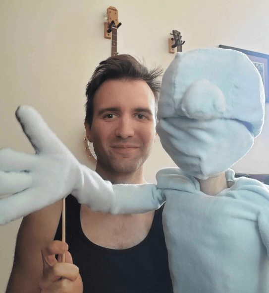 Kurt is operating a partially finished puppet. The puppet faces the camera. One of the puppet's hands is being raised by Kurt, moving it with a stick that goes into the wrist. Kurt looks like he's about to laugh.