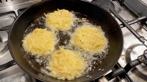 Four pale yellow potato latkes are sizzling in hot oil in a black frying pan on my stove