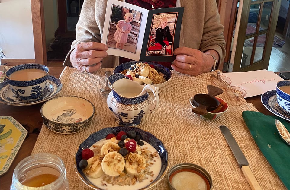 A woman sits at a breakfast table with oatmeal, fruit and coffee laid out. She holds a birthday card with two photos on either side.