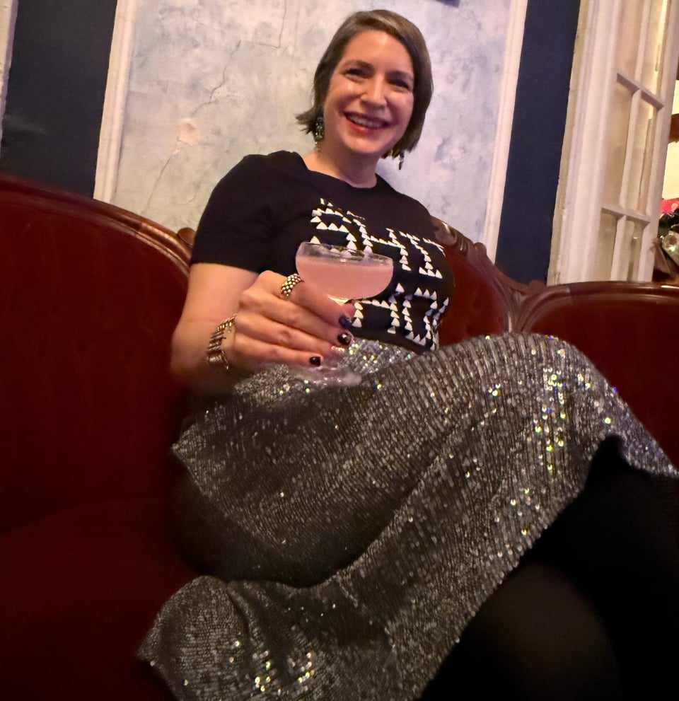Amanda Schuster wearing Punk Masters SHIT SHOW t-shirt and silver sequined skirt seated with cocktail on red velvet couch