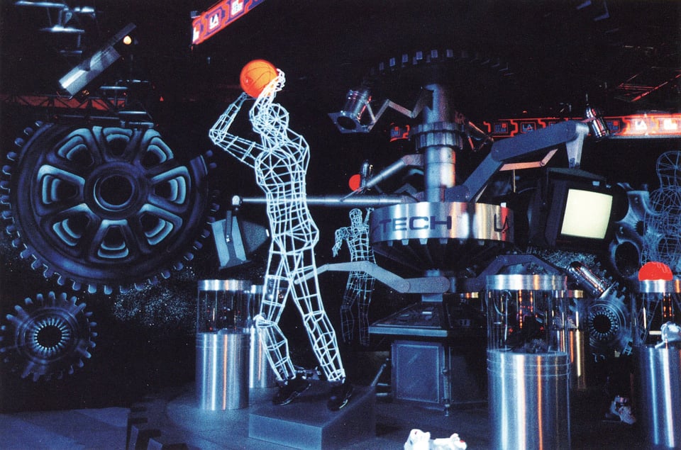 Within a Factory Pomo world of enormous metal gears and industrial structures, a light-up sculpture of a human being plays basketball.