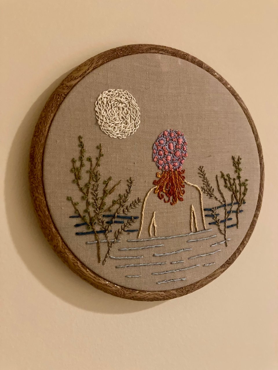 a hooped embroidery on a wall. it shows a woman's figure waist-deep in water, with red hair coming from beneath a floral bathing cap. branches frame either side of the water, and there's a full moon overhead.