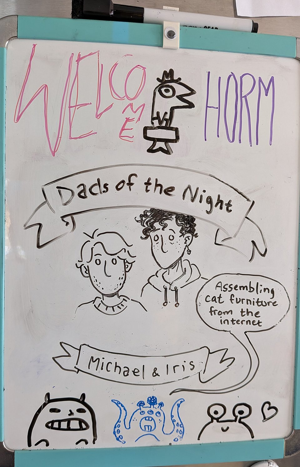 a photo of a fridge whiteboard. It says Welcome Horm along the top, and there are three weird little monster creatures along the bottom. In the middle is a banner that reads Dads of the Night, with a cartoony drawing of Michael and Iris, who both look somewhat haunted with empty eyes and no mouths. They received the Dads of the Night award, according to one of the whiteboard monsters, for "assembling cat furniture from the internet."