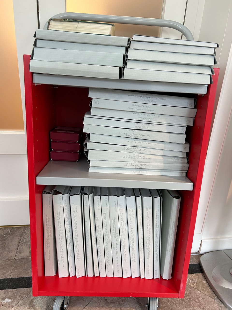 A red rolling set of shelves, full of grey folders that are each filled with archival documents.