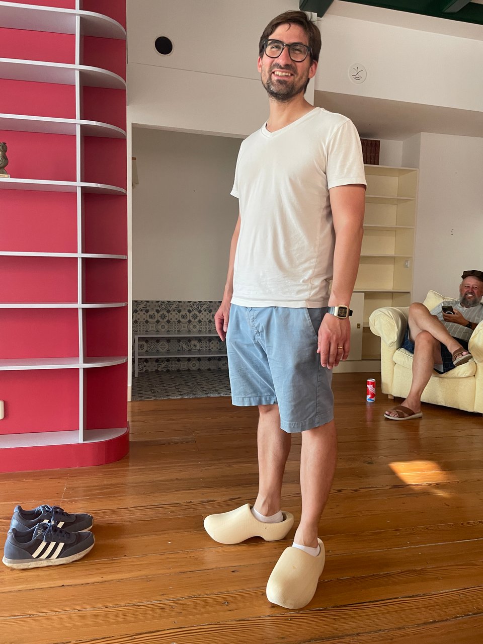 A smiling beared man stands in a room wearing blue shorts and a white t-shirt and large wooden Dutch clogs on his feet.
