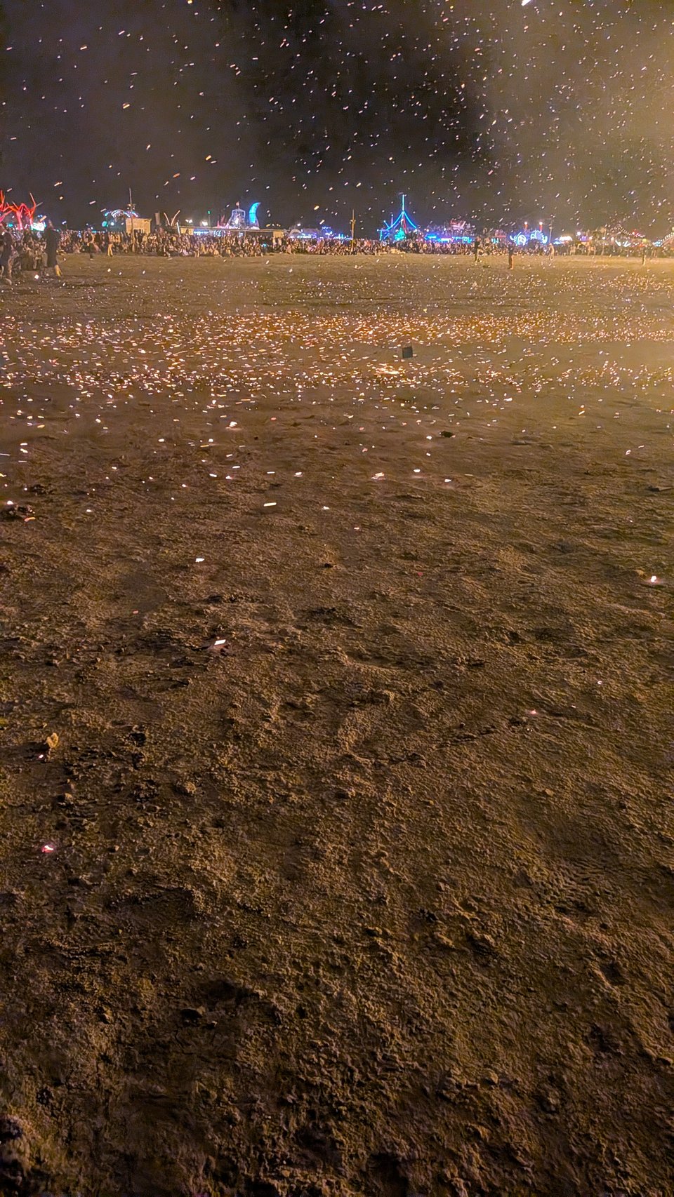 An image of the open playa, the Burning Man party in the background, as tiny bits of fire rain from the sky and scatter on the ground.
