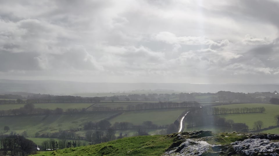 A cloudy day with the sun illuminating a road, a view down towards the coast from a hill