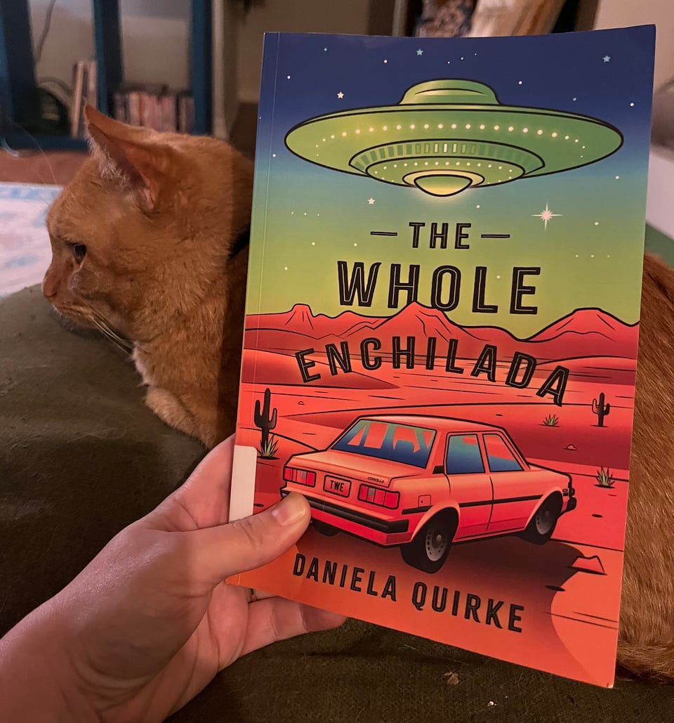 A white person's hand holds a paperback in front of an orange tabby cat on a green couch.