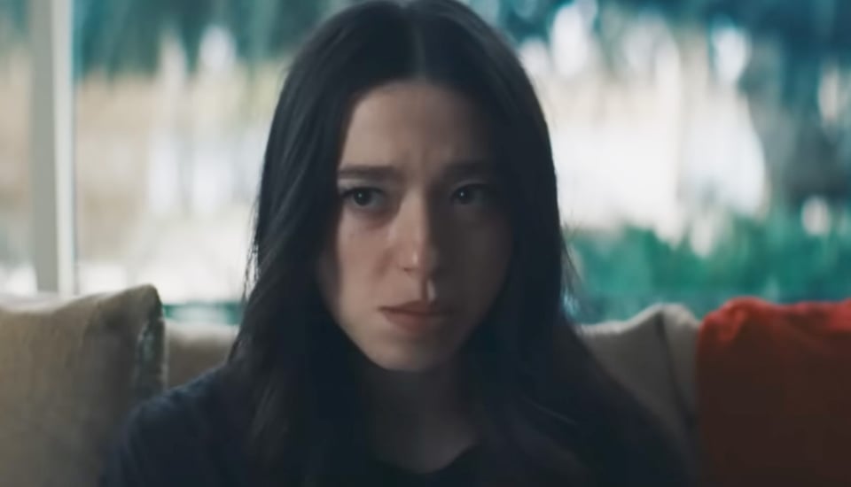 A young white woman with dark brown hair sitting on a beige sofa, wearing dark clothes, in front of a blurry green and white background. She has a sour expression on her face.