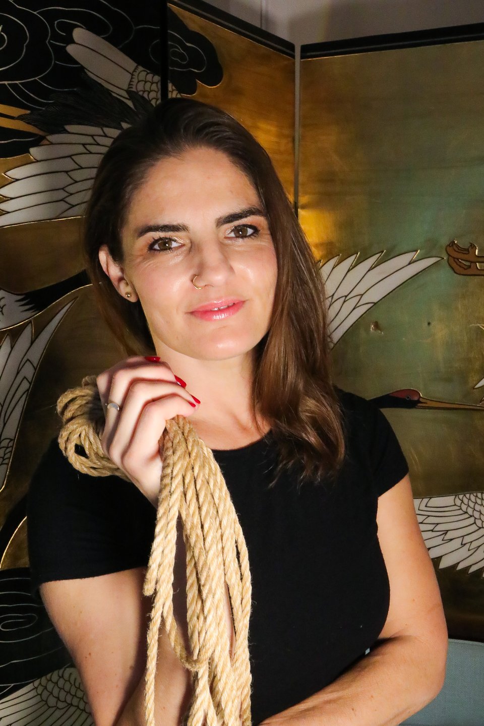 A portrait of a woman with shoulder-length brown hair and a nose ring, smiling softly at the camera. She wears a black t-shirt and holds a coil of thick, natural-fiber rope in her hand, showing off red nail polish. In the background stands an ornate gold and black Japanese folding screen depicting white cranes.