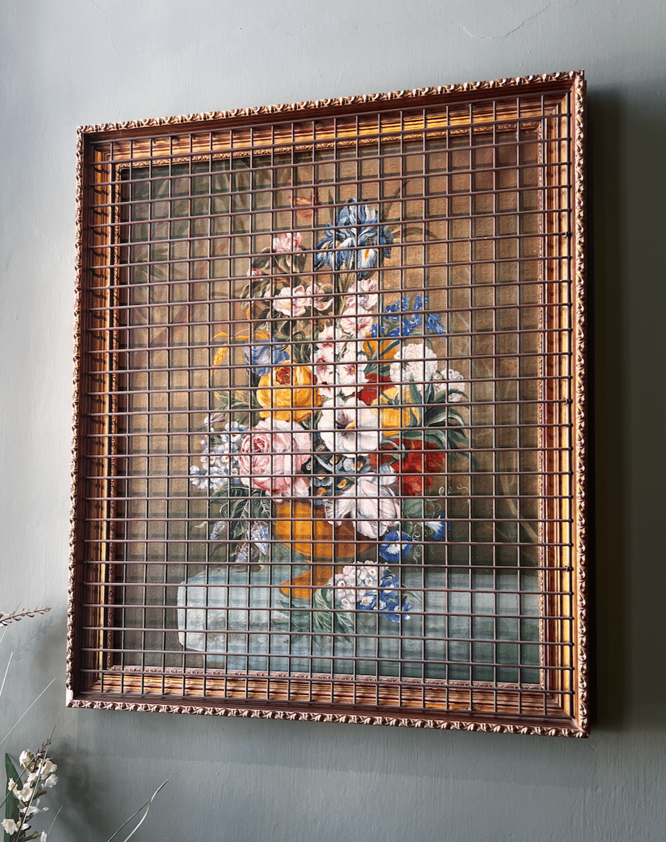 A classical painting of colorful flowers in a vase on a ledge, in a gold frame. The frame and painting are covered by a metal grate.