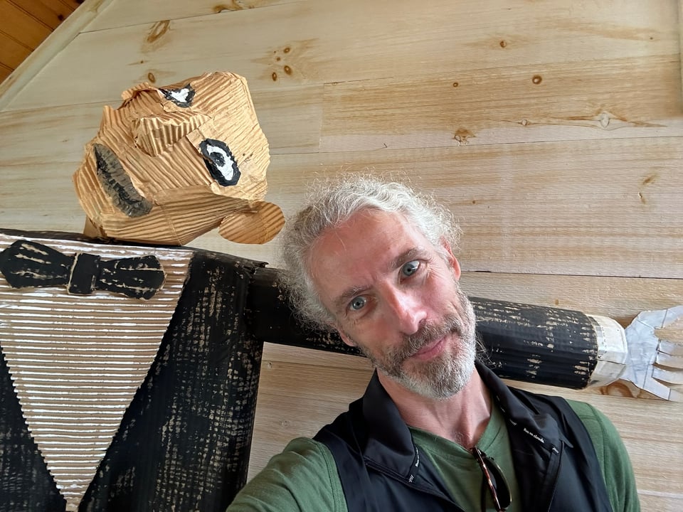 Photo of white man beside a textured cardboard sculpture of a man in a tuxedo. Both figures are tilting their heads toward each other