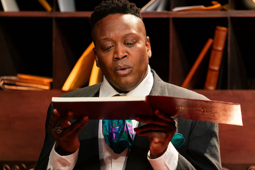 Tituss Burgess holds a book close to his face and makes a surprised face