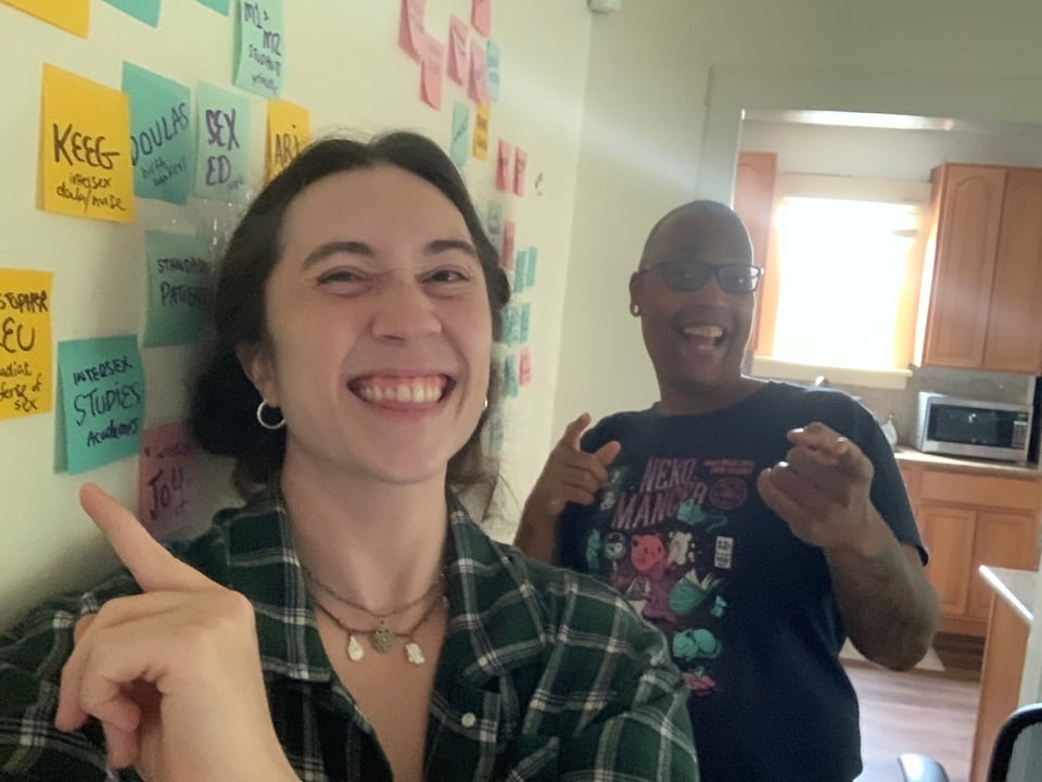 Hans and Saifa smiling in the kitchen, pointing to a wall of sticky notes from the planning retreat