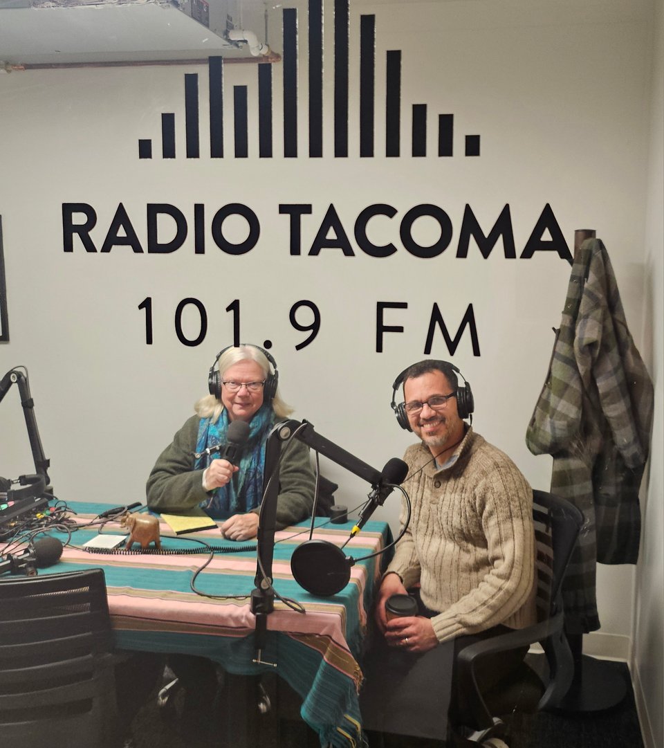 A woman and a man wearing headsets sit together at microphoones in a recording studio